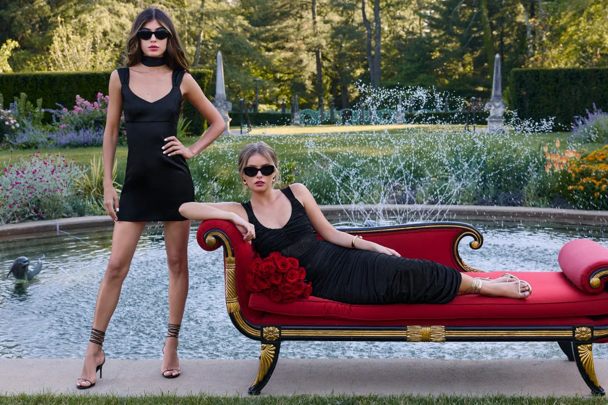 A lifestyle shot of two young women in black dresses and sunglasses posed by a red velvet settee beside an ornamental garden pool.