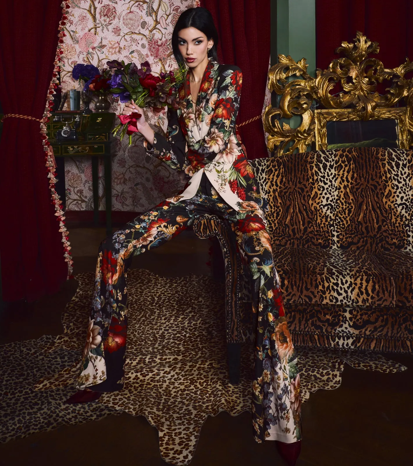 A lifestyle shot of a woman wearing a black floral-print blazer and matching wide-leg trousers in an ornate, jewel-toned interior setting with luxe furnishings.