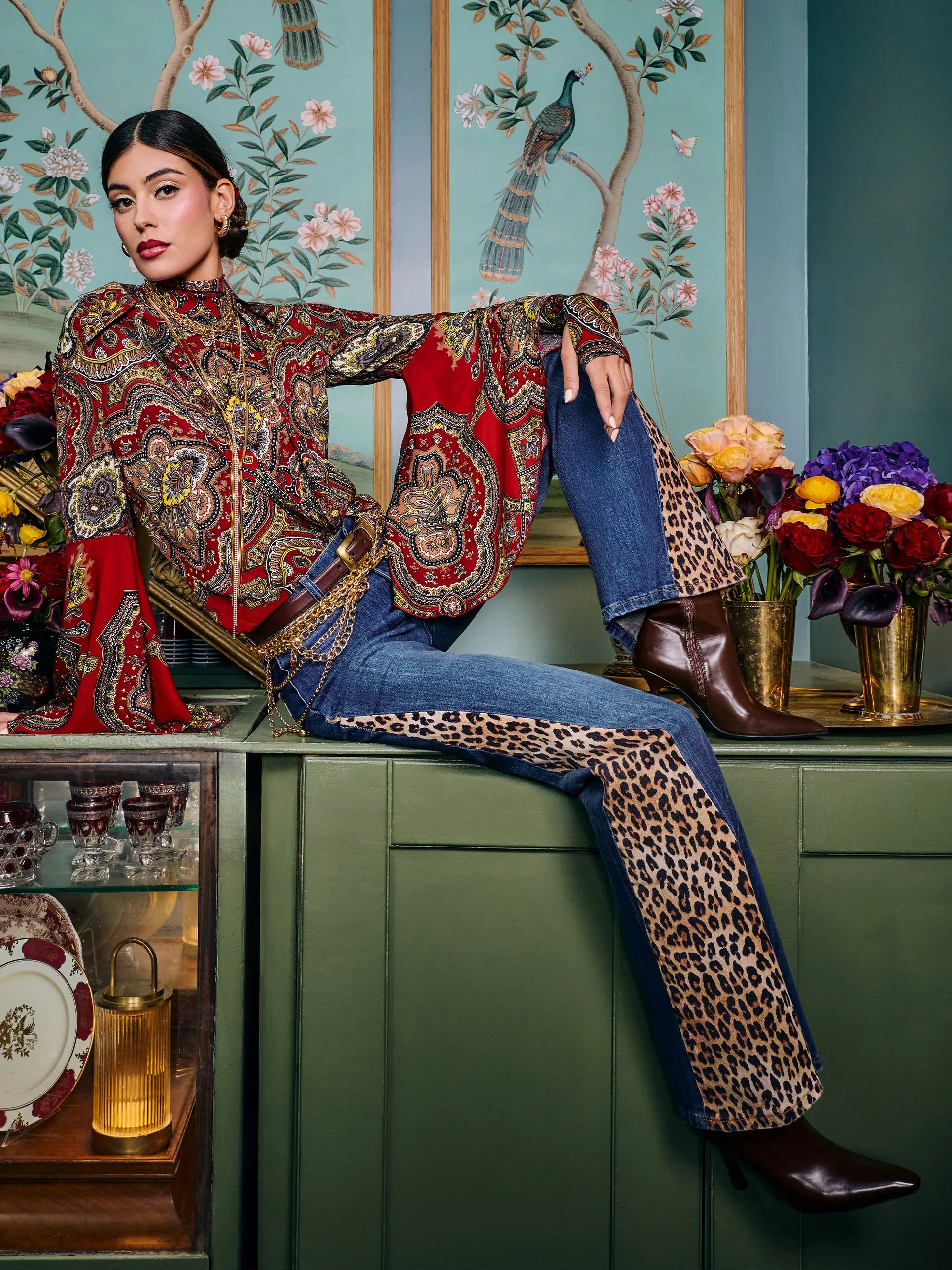 A lifestyle shot of a woman wearing a red and gold patterned jacket and leopard-print denim, seated against an ornate turquoise wallpapered backdrop with botanical motifs and brass accents.