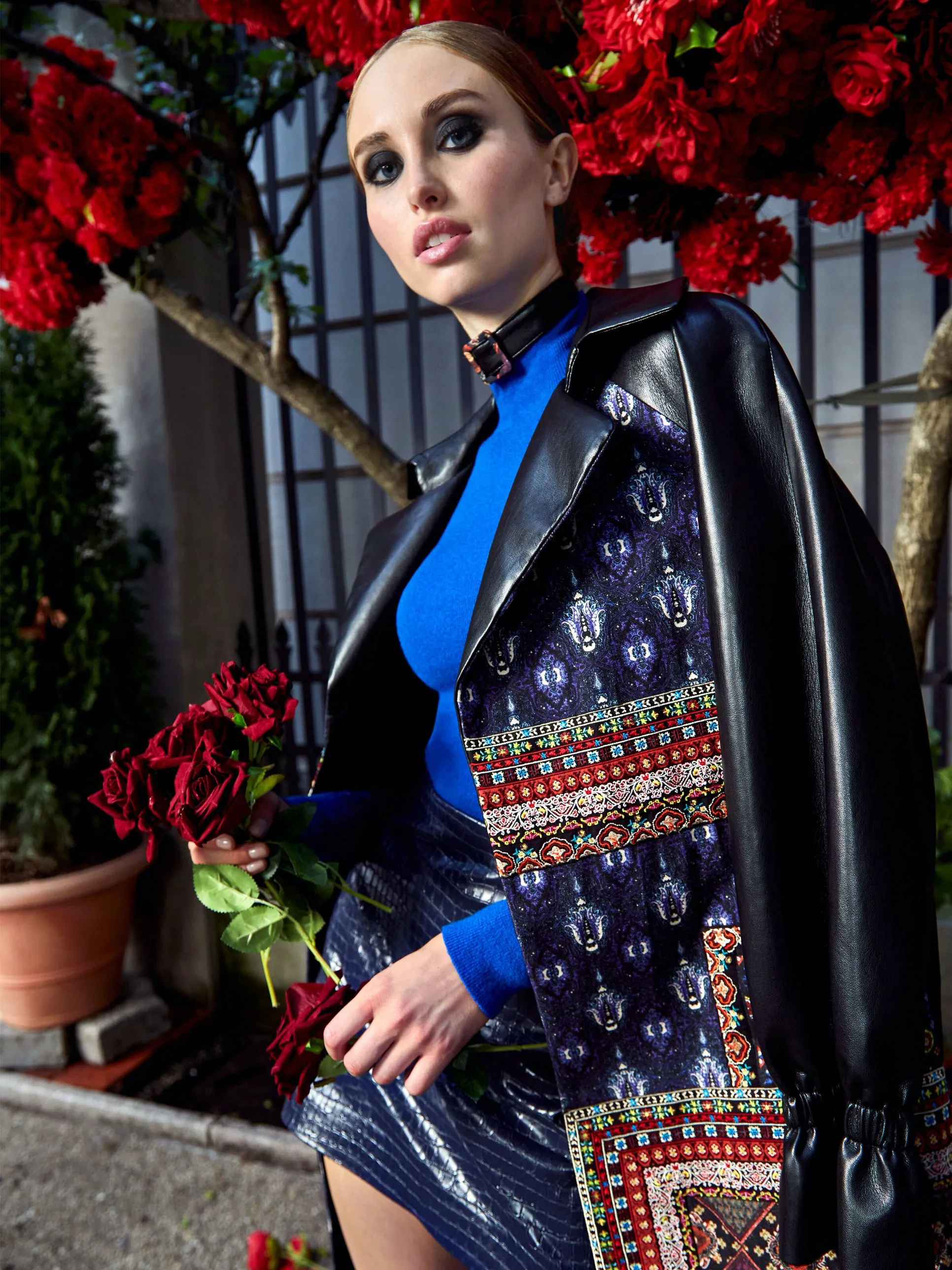 A lifestyle shot of a woman wearing a black leather jacket and blue knit top over a patterned navy dress with an ornate woven belt, holding red roses among blooming flowers.