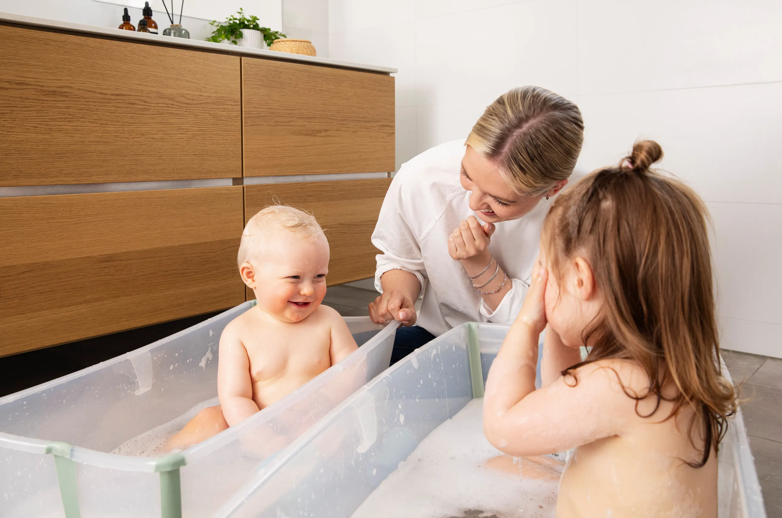 A lifestyle shot of a smiling woman in a white shirt leaning over two children bathing in transparent green Stokke FlexiBath tubs in a bright modern bathroom with warm wood cabinetry.