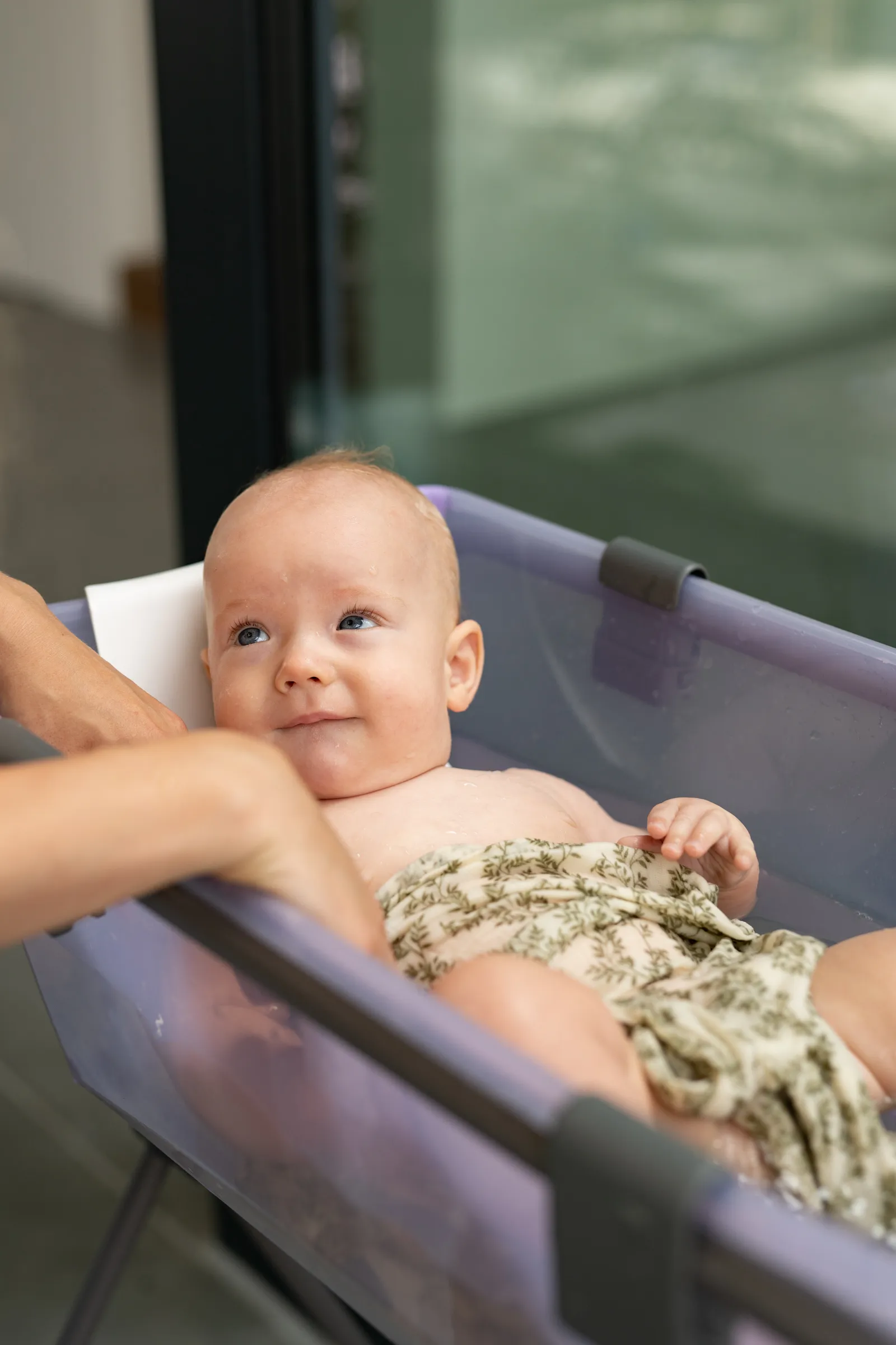 A lifestyle shot of an infant in a purple bathing tub receiving a bath in soft natural light.