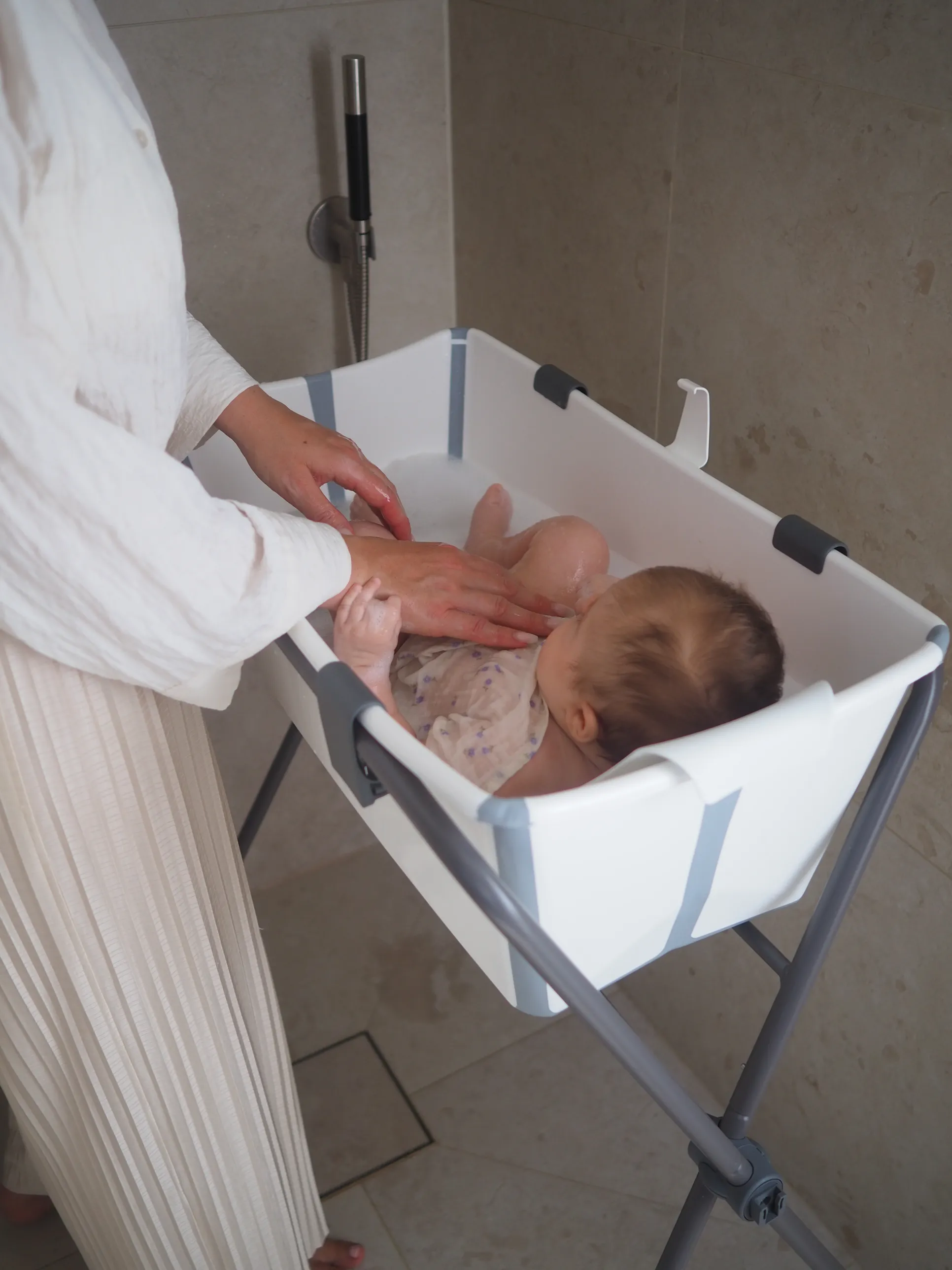 A lifestyle shot of a caregiver bathing a newborn infant in a white portable baby bath basin in a modern bathroom setting.