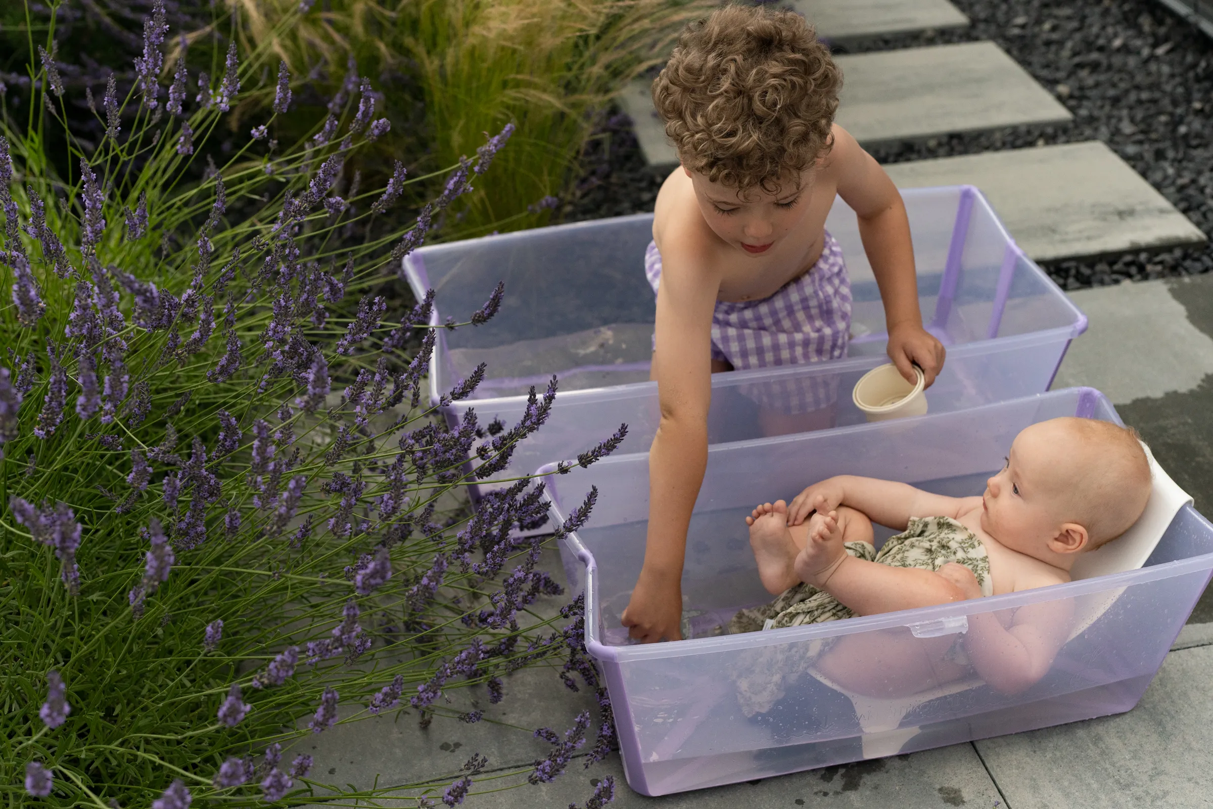 A lifestyle shot of two children bathing in purple plastic tubs surrounded by lavender plants in a sunny garden setting.