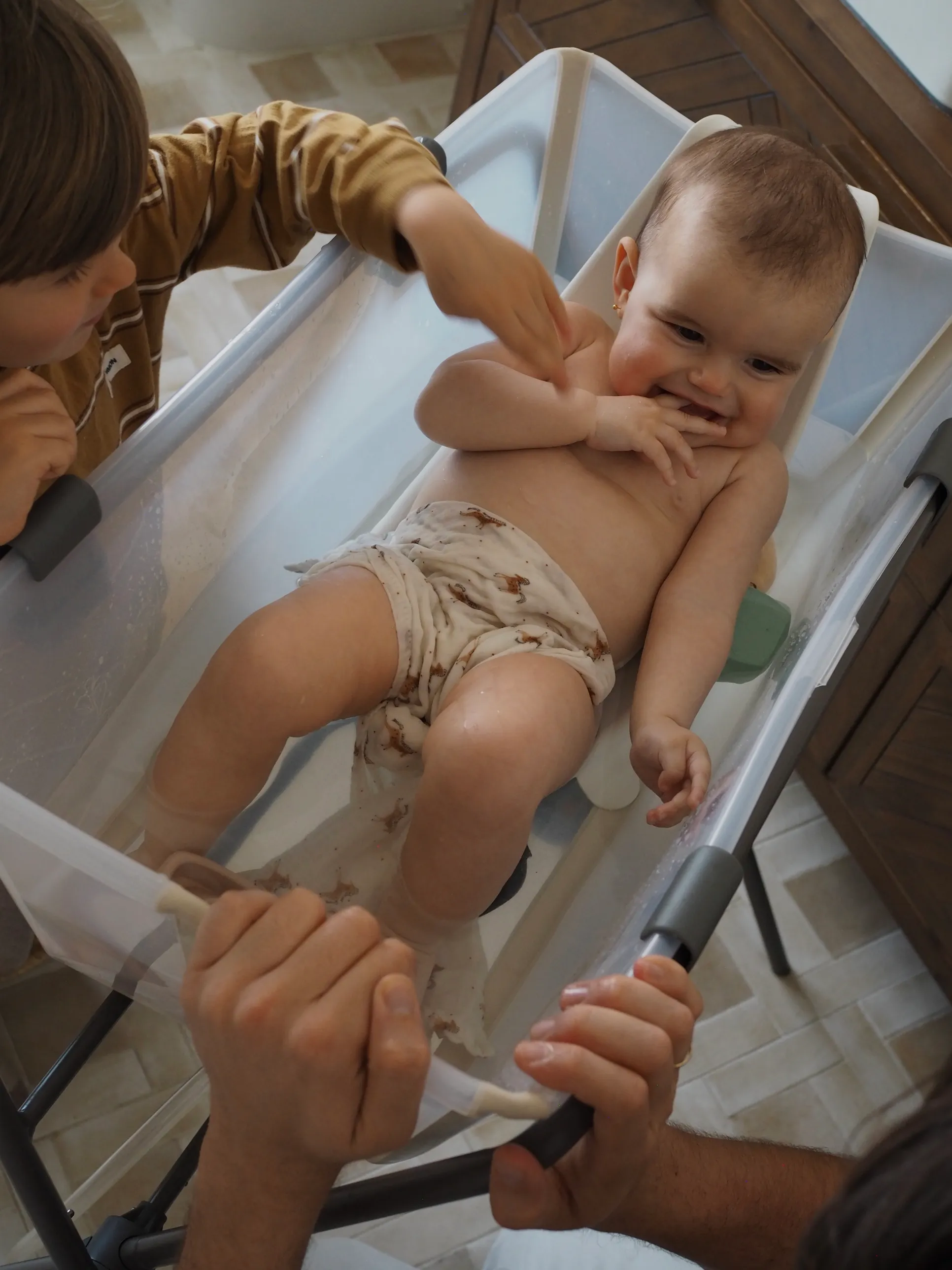 A lifestyle shot of a mother and father bathing their toddler in a white baby bath, with warm water and the child smiling contentedly.