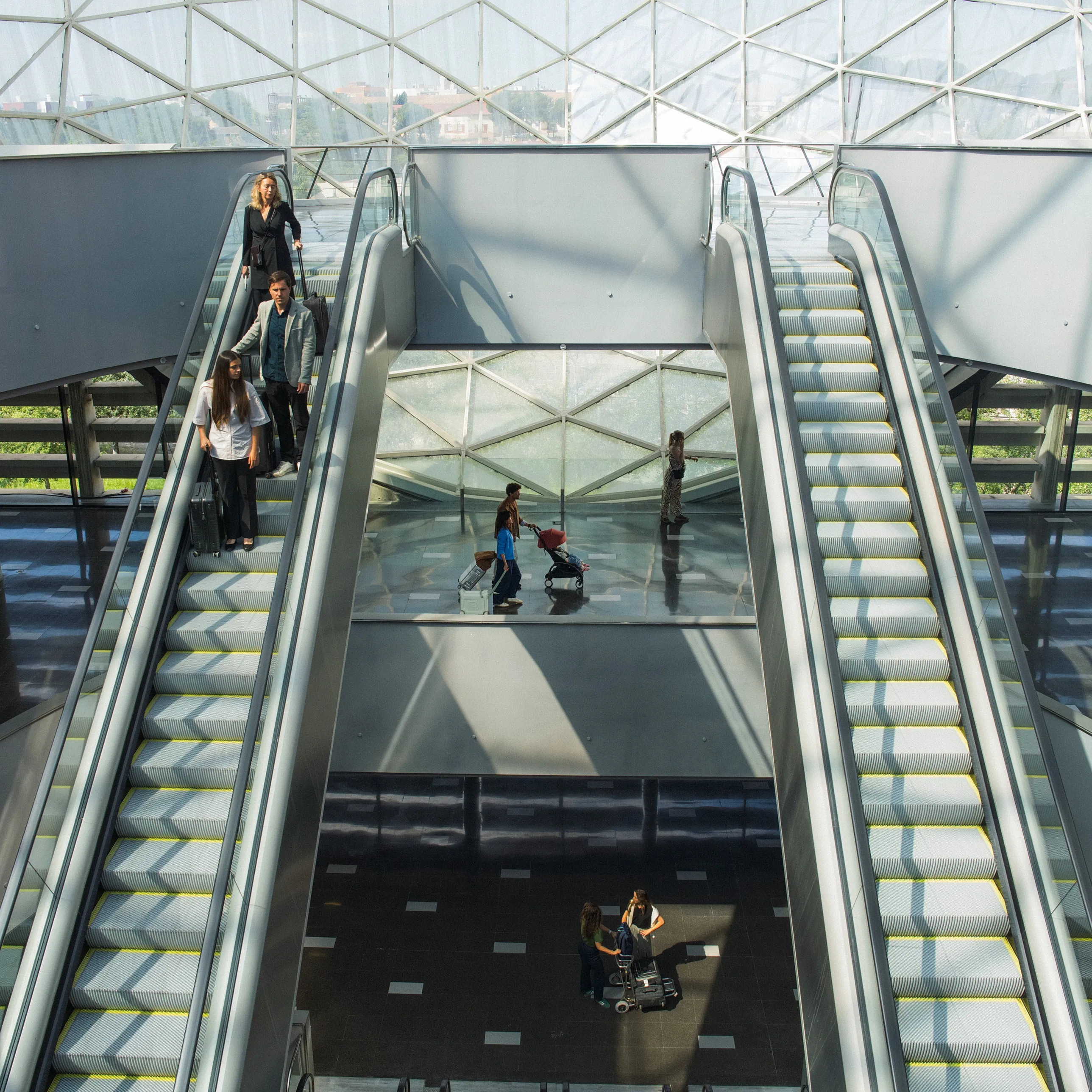 A lifestyle shot of a family with a Stokke YOYO5 stroller crossing a glass-roofed terminal with escalators and travelers with luggage in soft daylight.