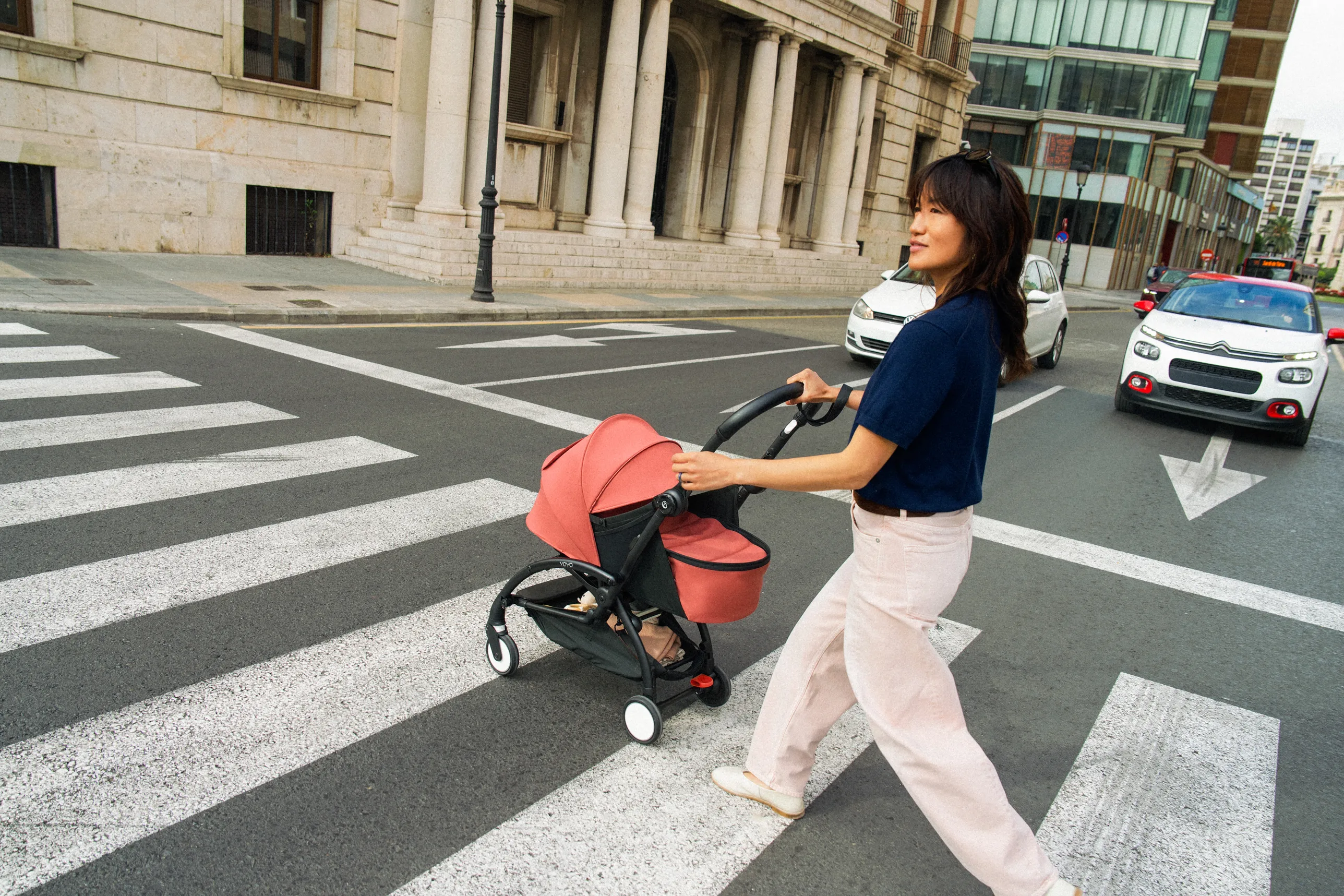 A lifestyle shot of a woman pushing a red and black stroller across an urban street crosswalk, surrounded by buildings and traffic.