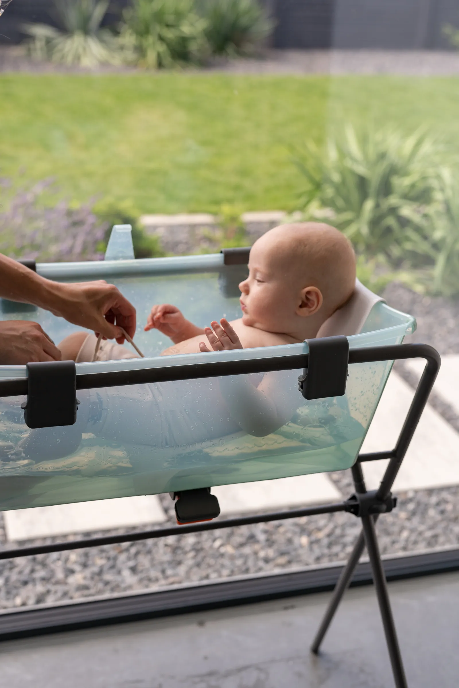 A lifestyle shot of a baby bathing in a blue portable bath with an adult's hands nearby in a sunny outdoor garden setting.
