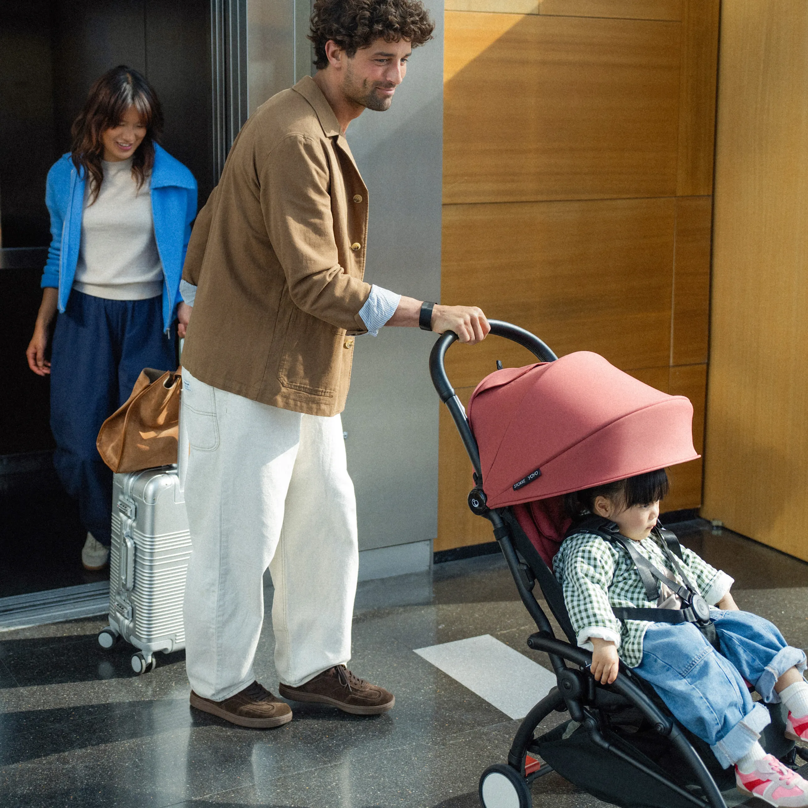 A lifestyle shot of a man in a brown jacket and cream trousers pushing a Stokke YOYO5 stroller in berry canopy through a lobby with a woman and rolling suitcase behind him.
