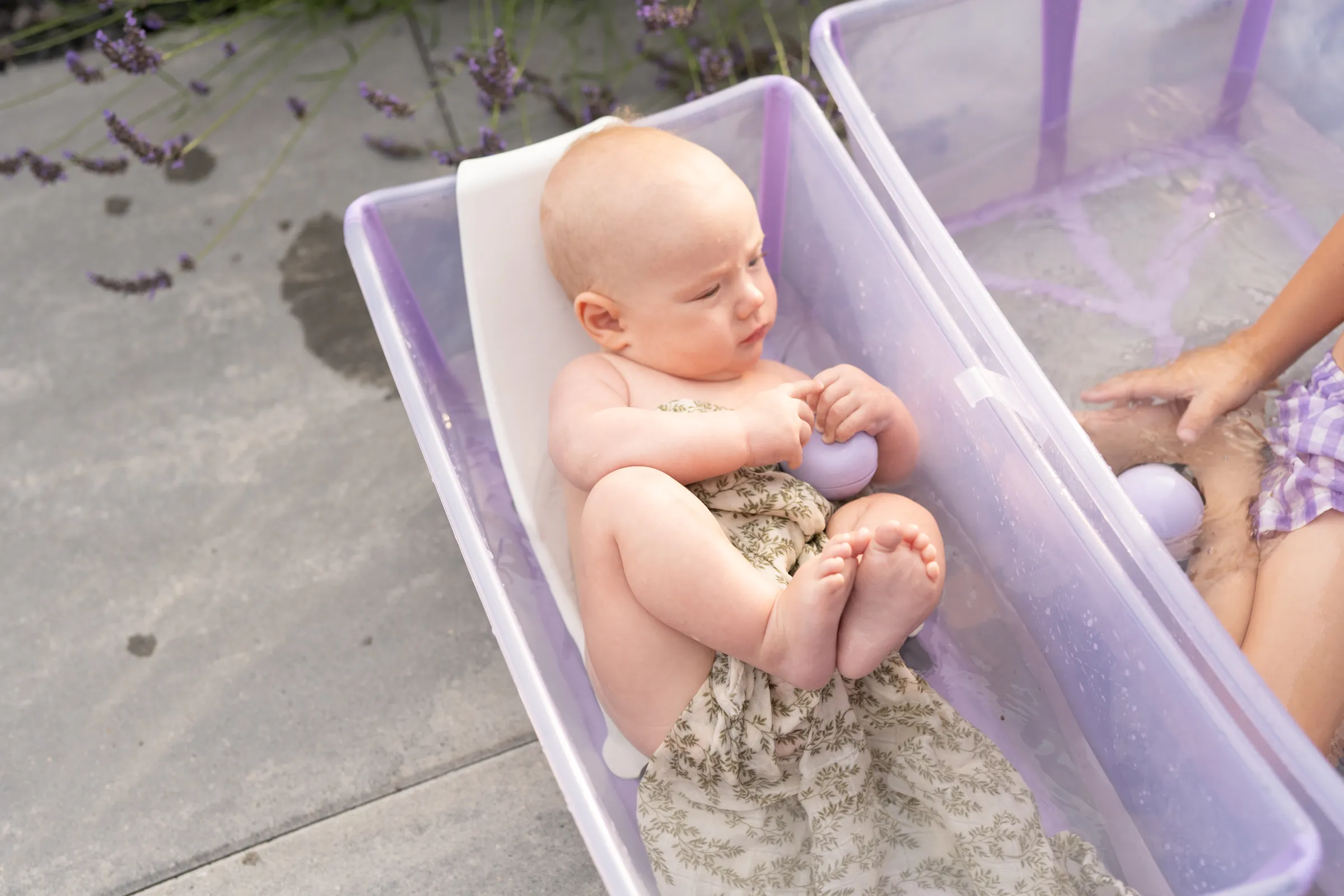 A lifestyle shot of a baby playing in a purple bath tub filled with sand and lavender flowers outdoors.
