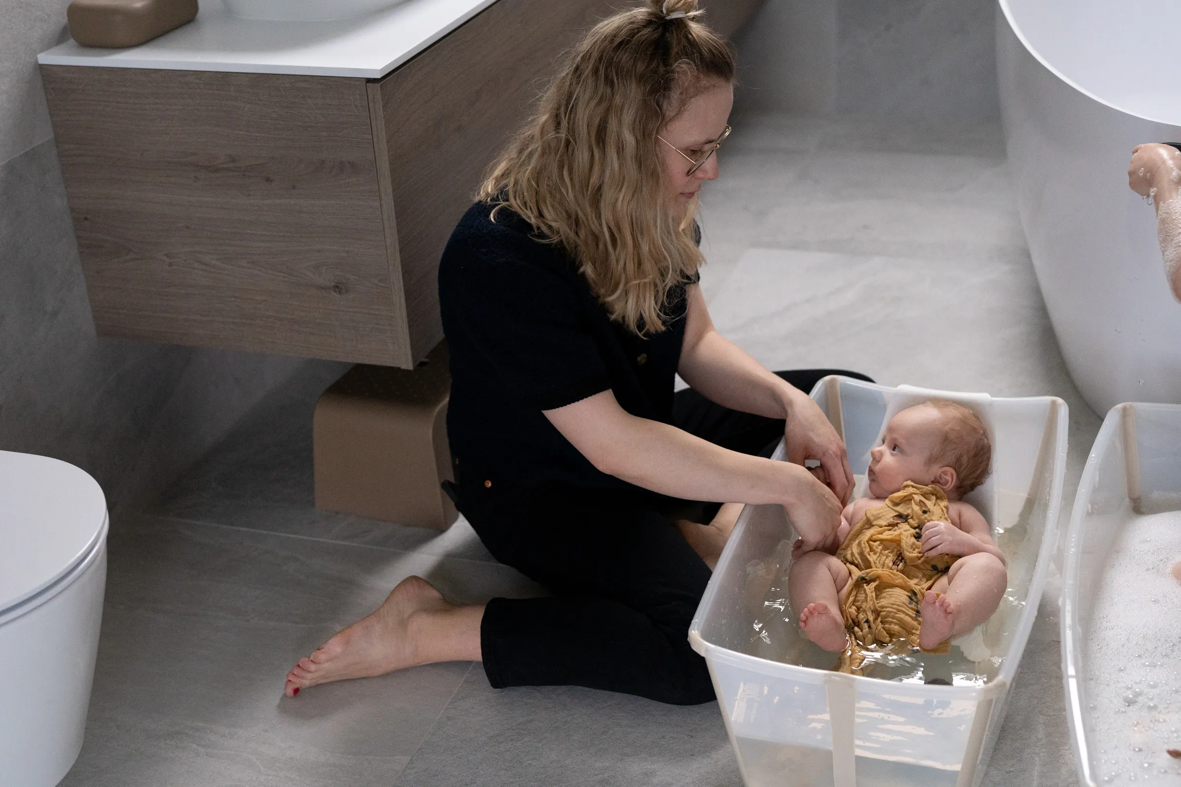 A lifestyle shot of a woman bathing a baby in a white basin on a modern bathroom floor, with minimalist vanity and toilet in the background.
