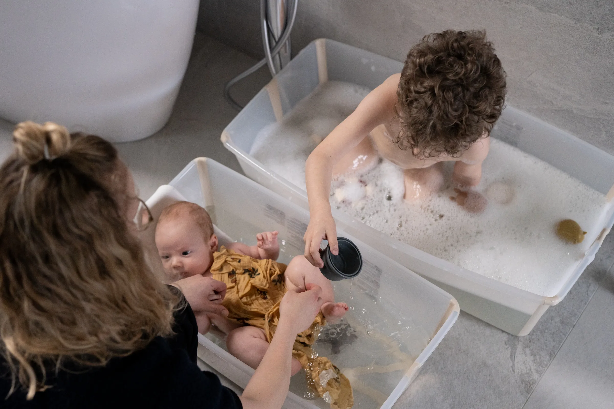 A lifestyle shot of a woman bathing a baby in a translucent FlexiBath tub while a curly-haired toddler plays in foamy water in a second tub in a bright bathroom.