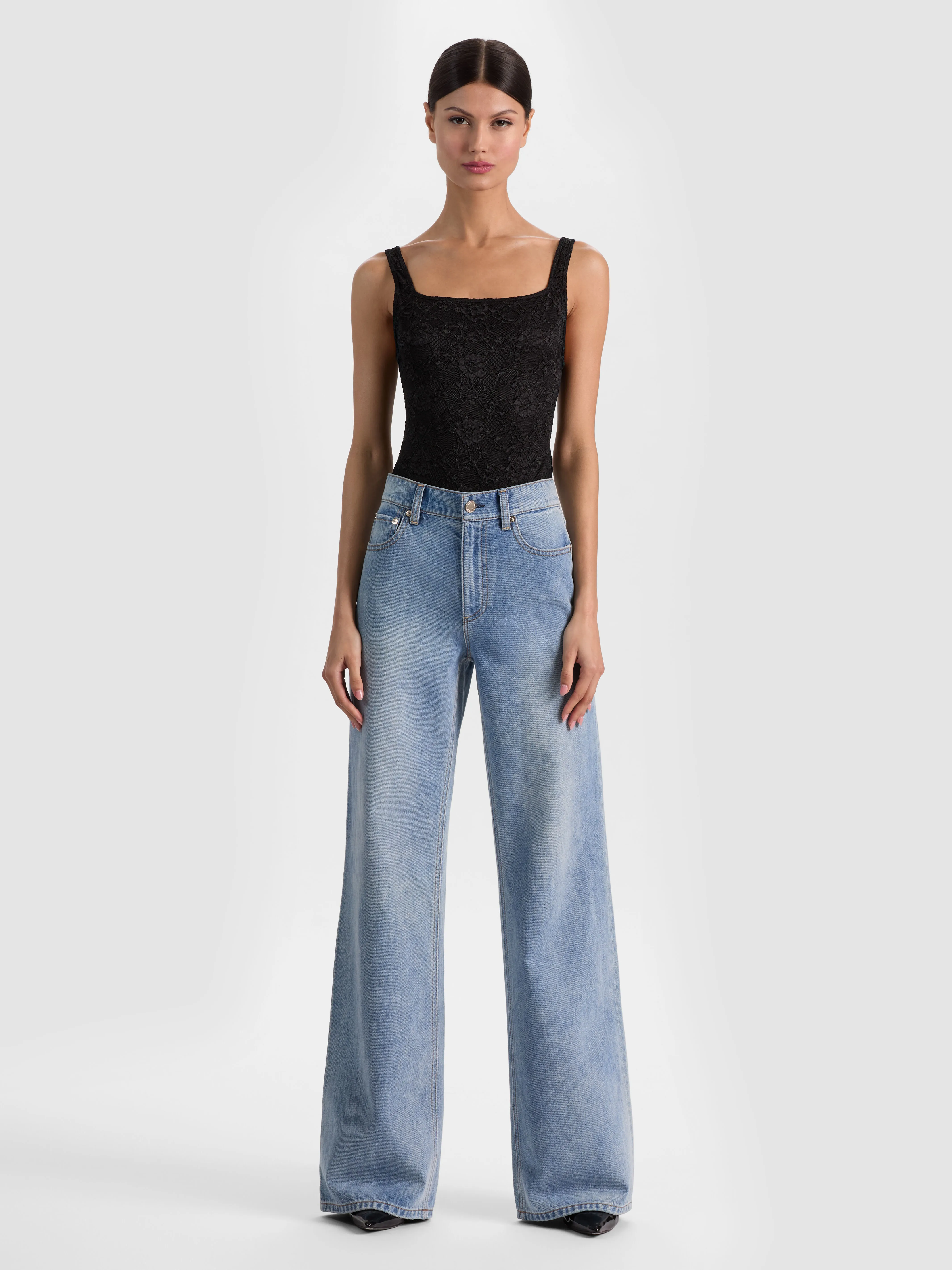 A full-body head-to-toe shot of a model wearing the ALICE + OLIVIA Annika Lace Bodysuit tucked into wide-leg jeans, styled for a sleek and sophisticated work or night-out look.