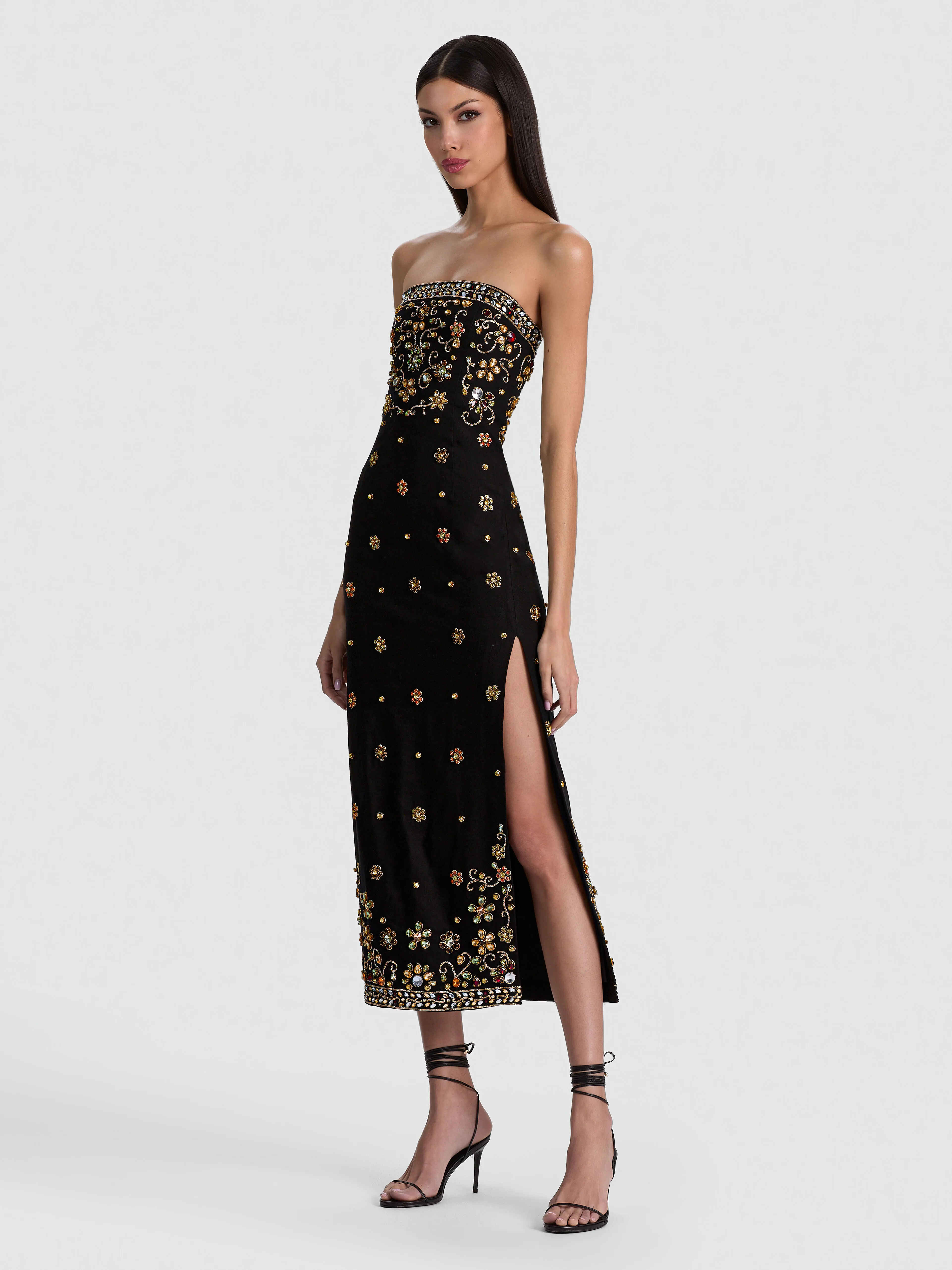 A model wearing the ALICE + OLIVIA Nina Embellished Midi Dress in a strapless black silhouette with intricate multicolored crystal embroidery, styled with statement earrings and strappy heels for a formal evening event.