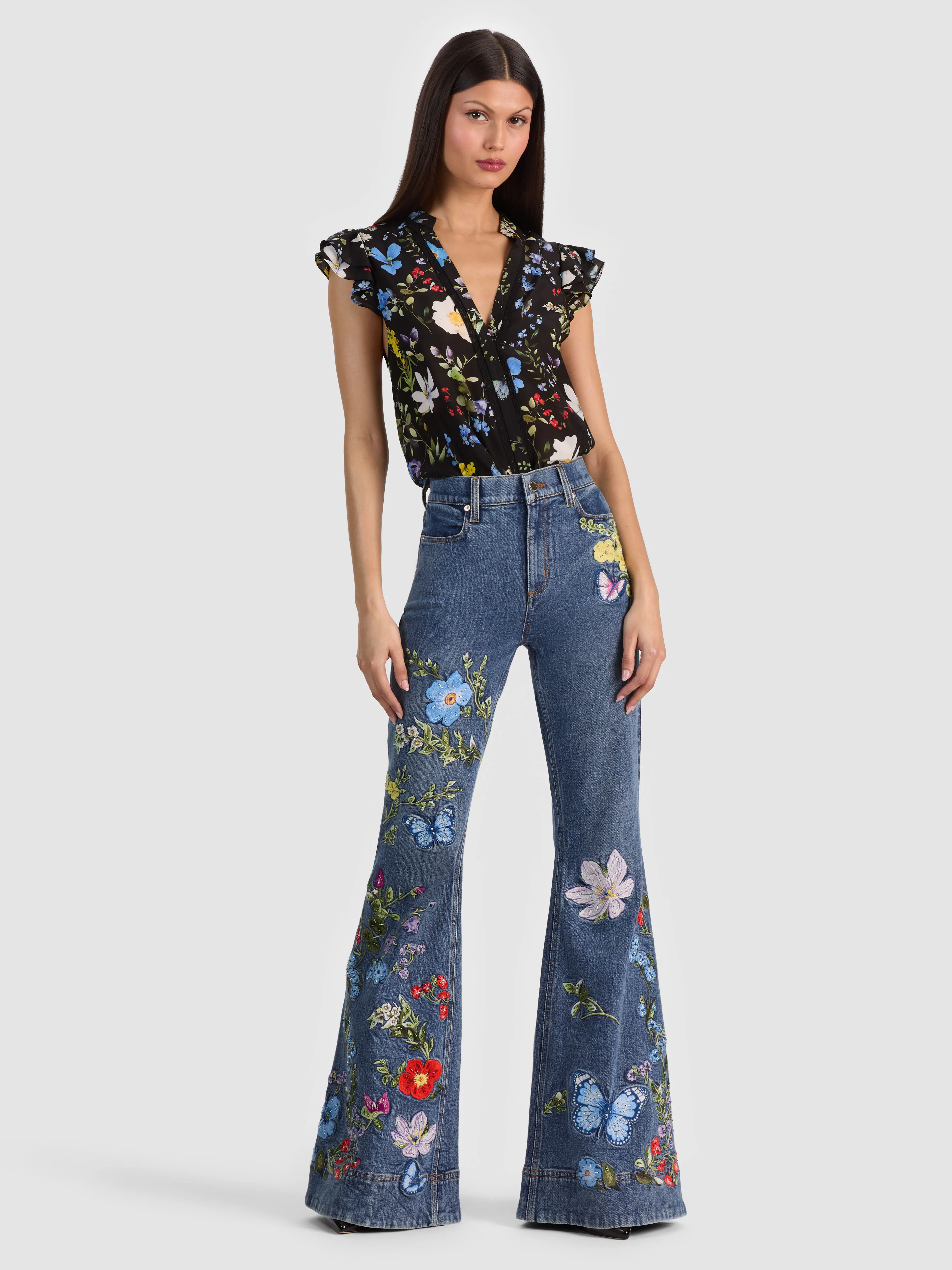 Full-body head-to-toe shot of a model wearing the ALICE + OLIVIA Beautiful Embroidered Jean in Brooklyn Blue/Multi, styled with a silk blouse tucked in, showing the model's face and complete outfit.