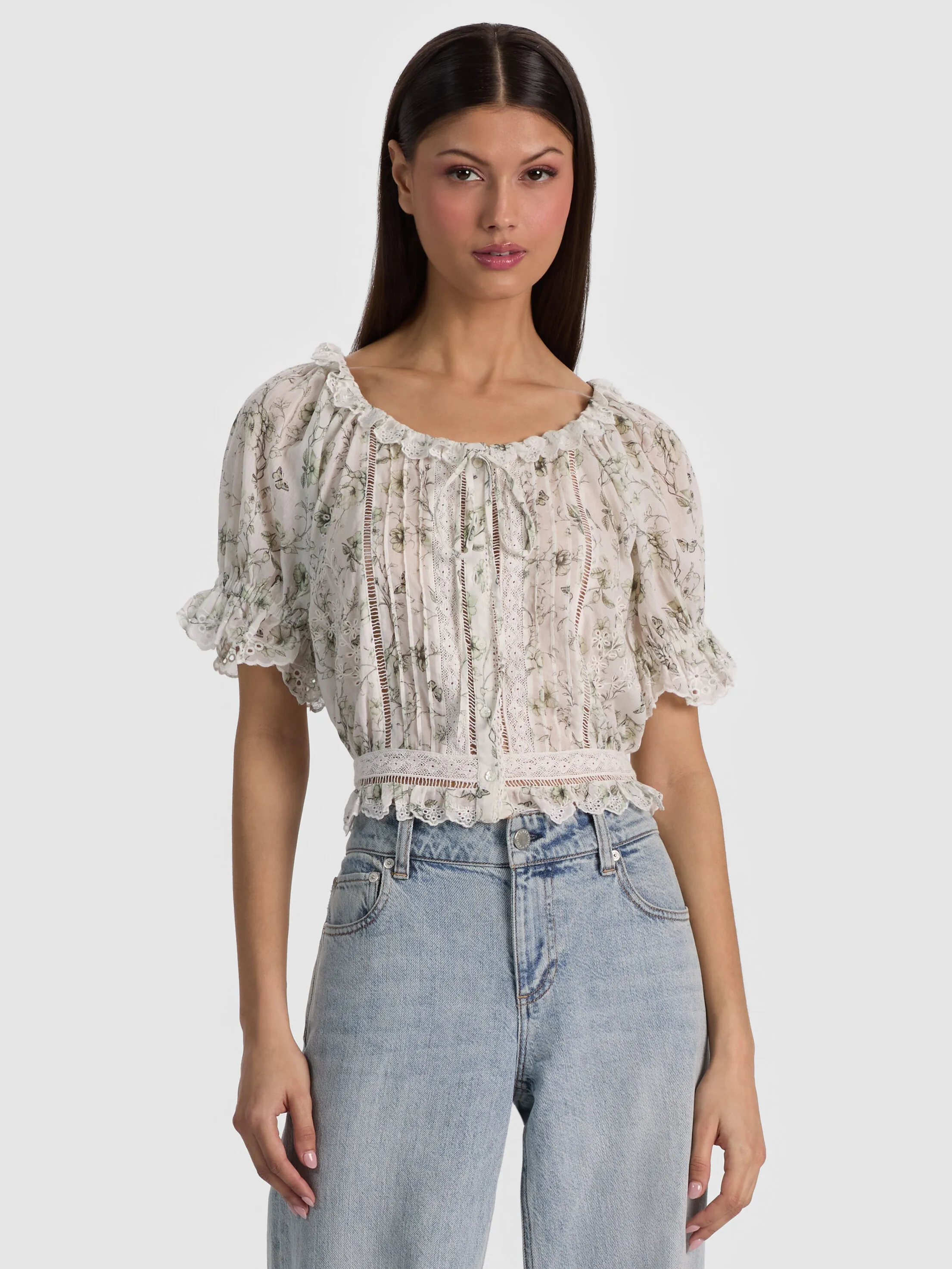 A woman wearing the McLaine Puff Sleeve Blouse in sage green floral cotton voile, walking through a bright city neighborhood, blouse catching a warm summer breeze.