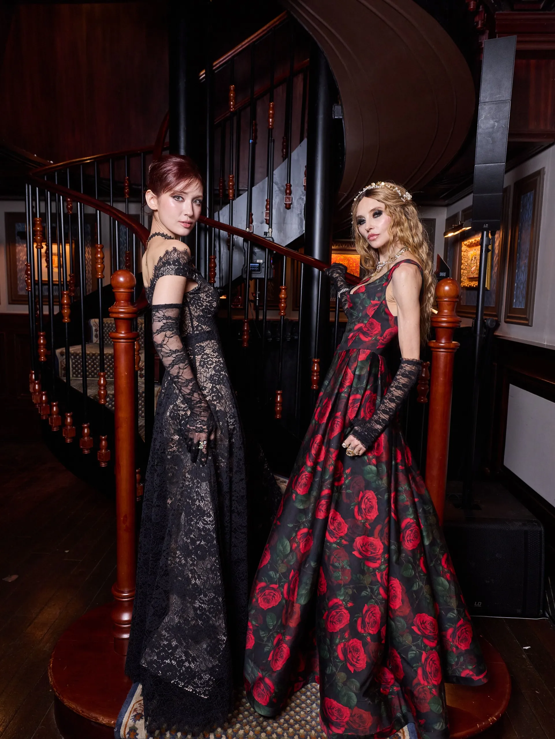 A lifestyle shot of two women in black eveningwear posed on a vibrant pink velvet carpet surrounded by flowers, desserts, and ornate furnishings against red curtains and blue velvet seating.