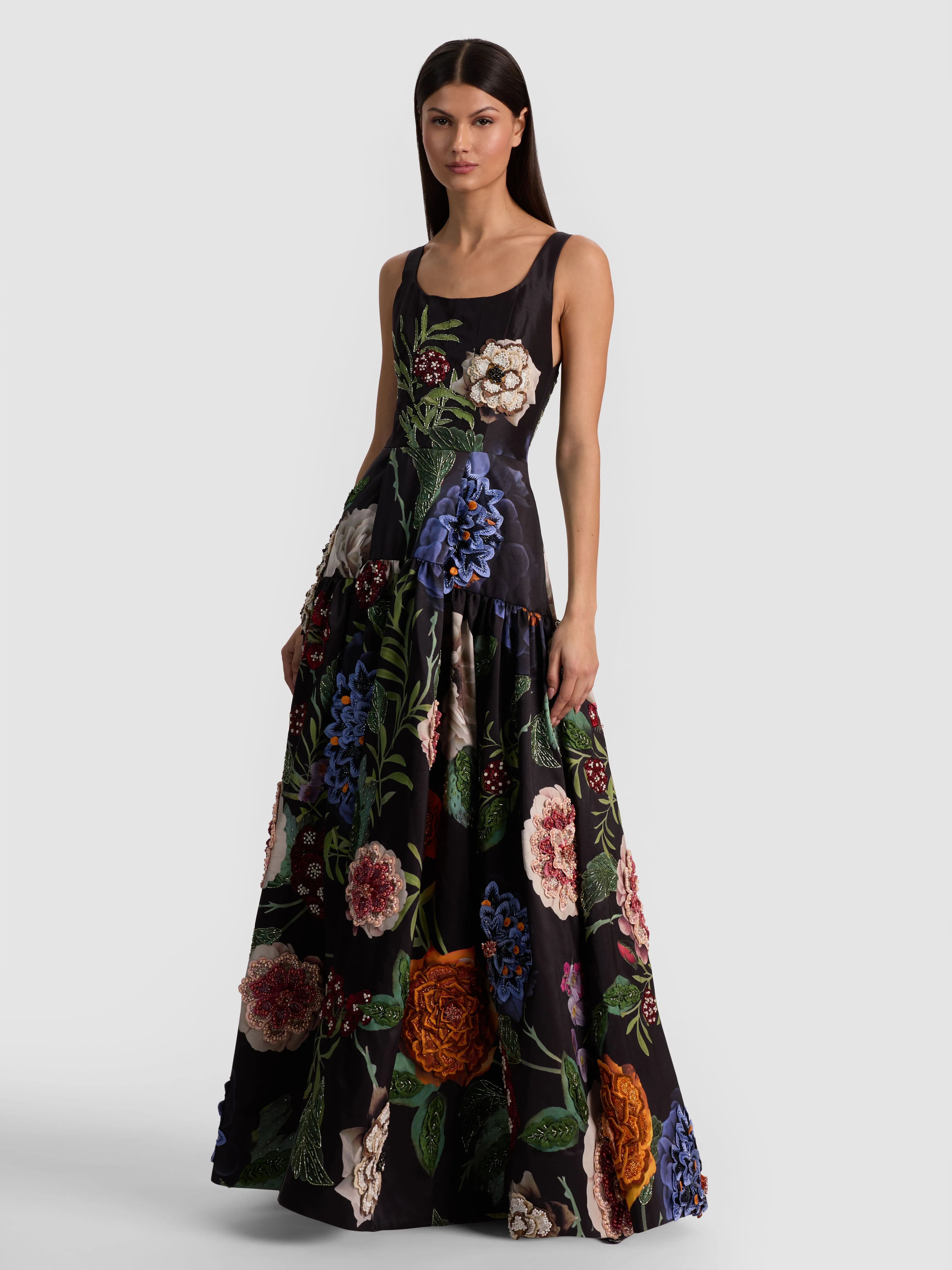 A model wearing the Maxine Embellished Gown in black, styled glamorously with strappy metallic heels, sparkling earrings, and a clutch for a formal evening event.
