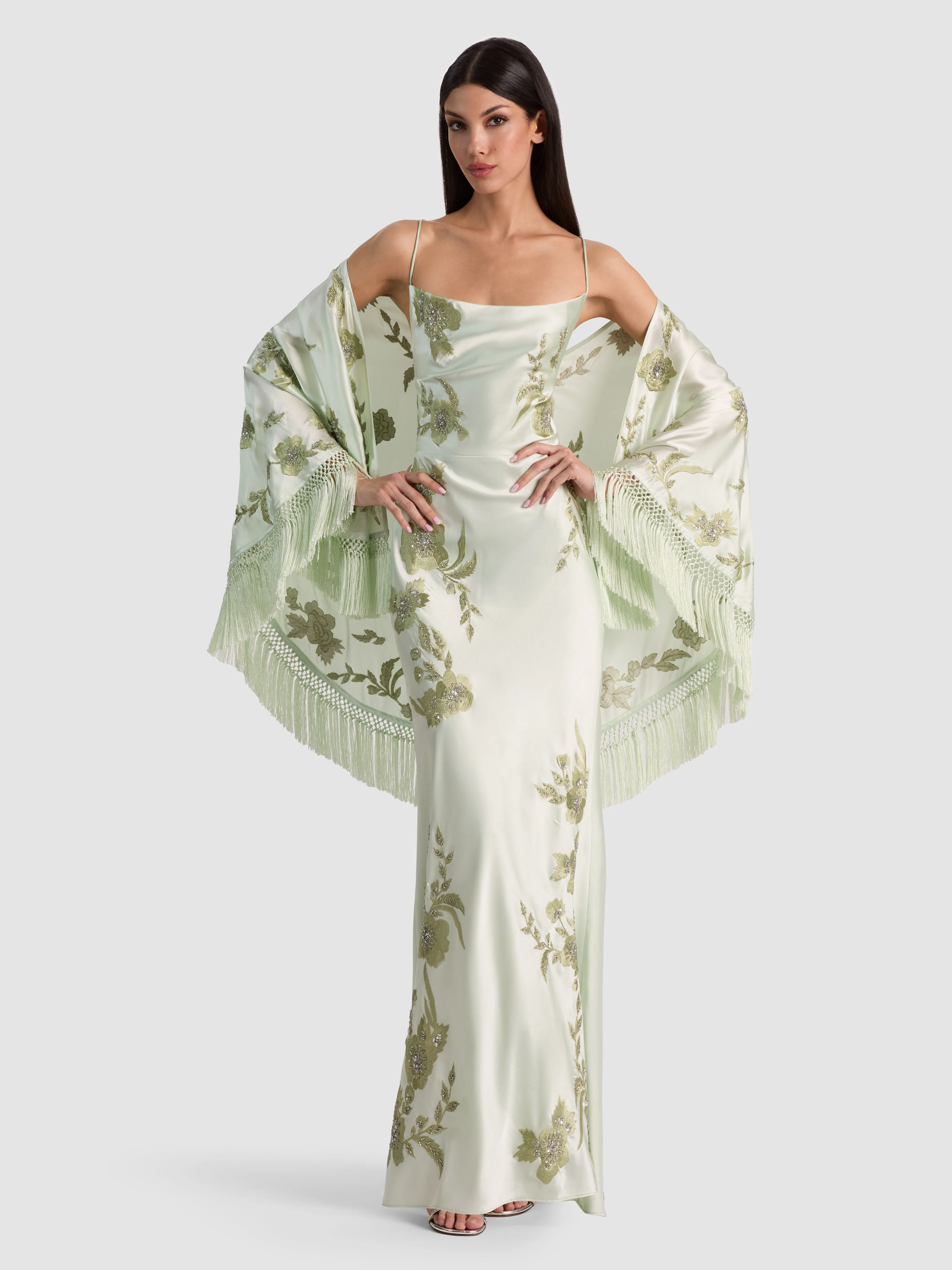 A model wearing the ALICE + OLIVIA Marlene Shawl Scarf in Green Lily/Multi, a hand-embellished crystal shawl with fringe trim draped over the shoulders over a dress.