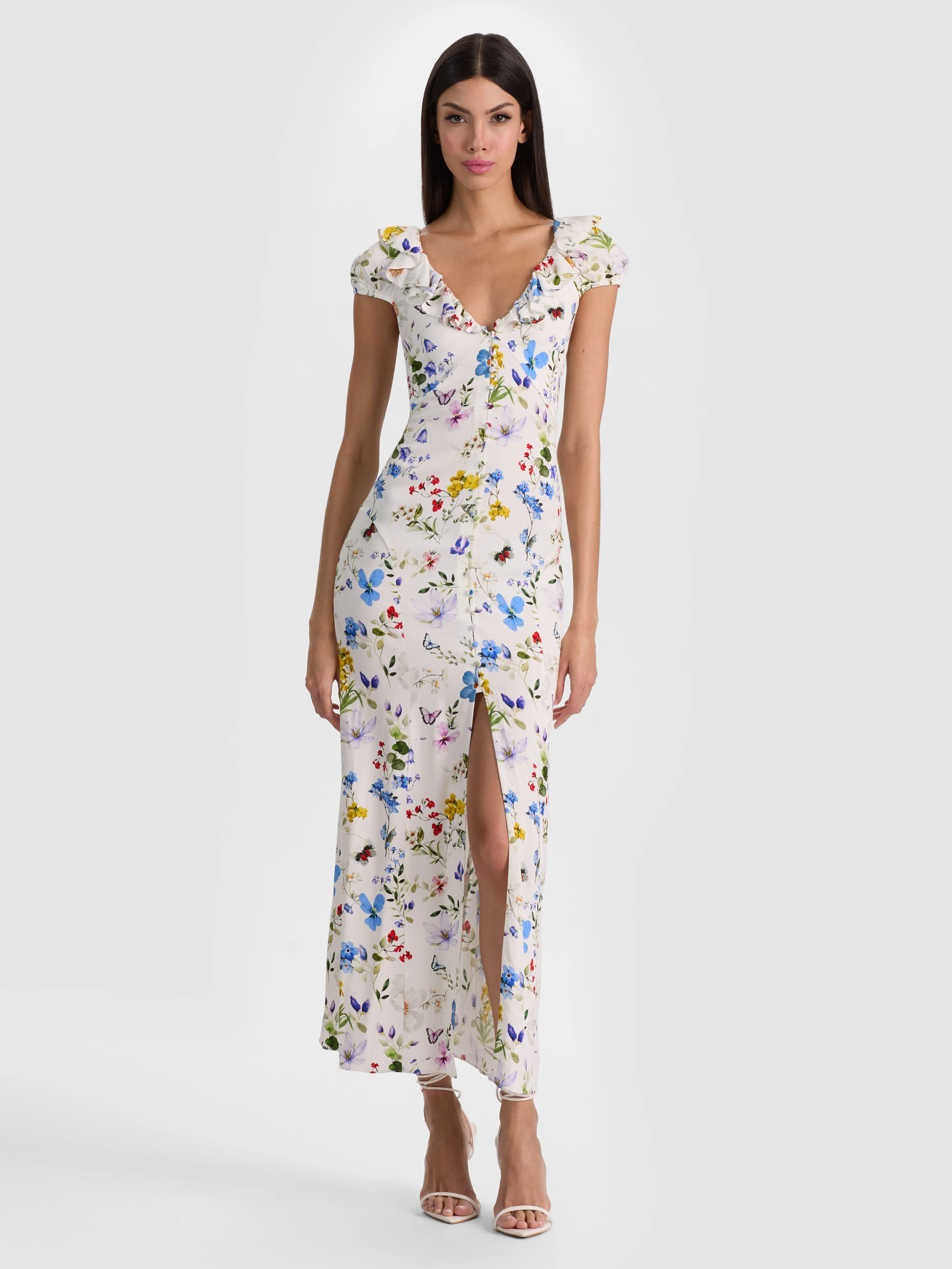 A lifestyle shot of a woman wearing the TARA MIDI SLIP DRESS — floral printed viscose crepe with a deep V neckline, ruffle detailing, and a back waist tie — styled with strappy heels for an elegant evening look.