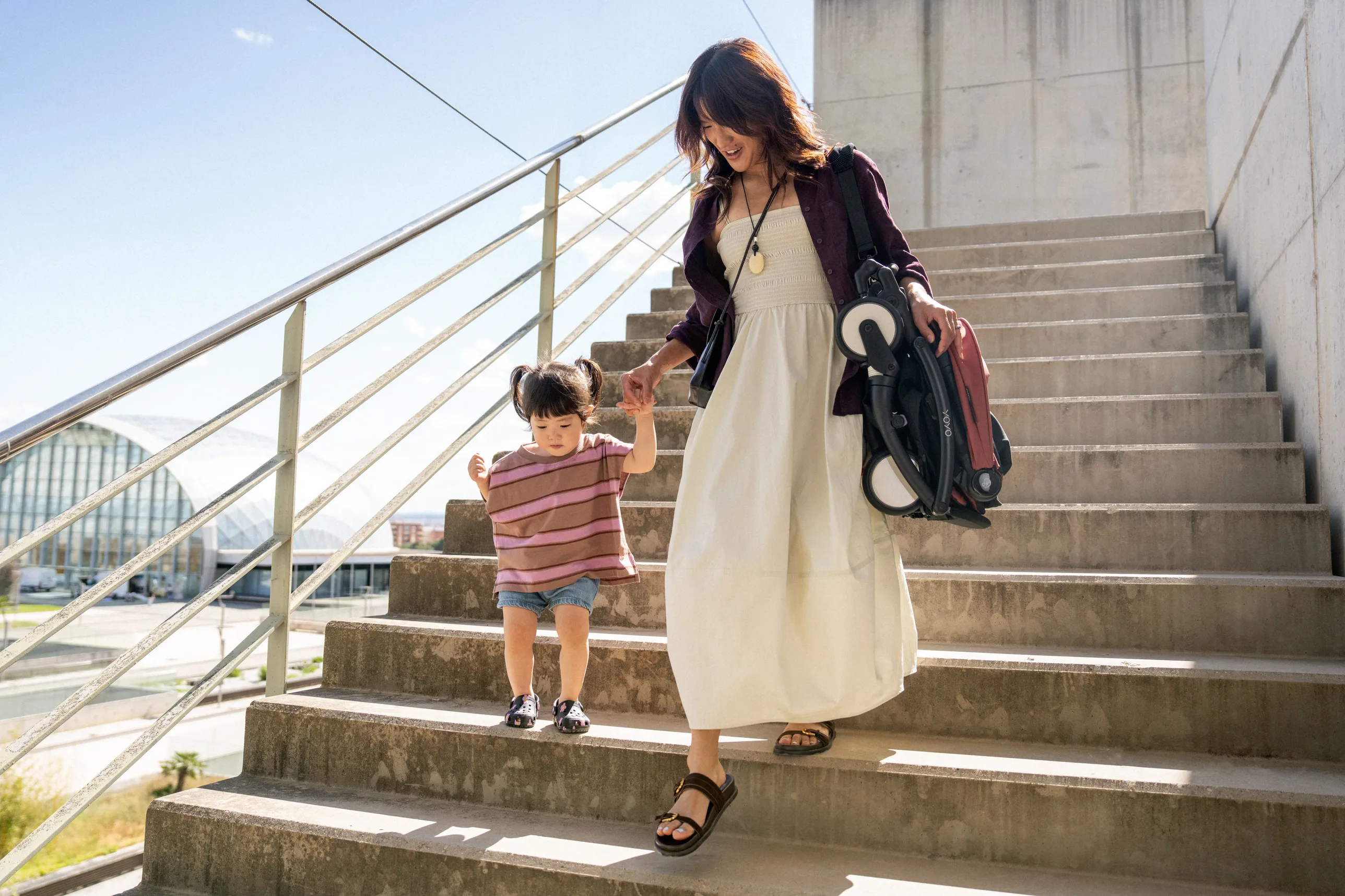 A lifestyle shot of a parent pushing a lightweight YOYO⁵ compact stroller with a black frame on a sunny urban street, showcasing its compact design and easy one-hand steering.