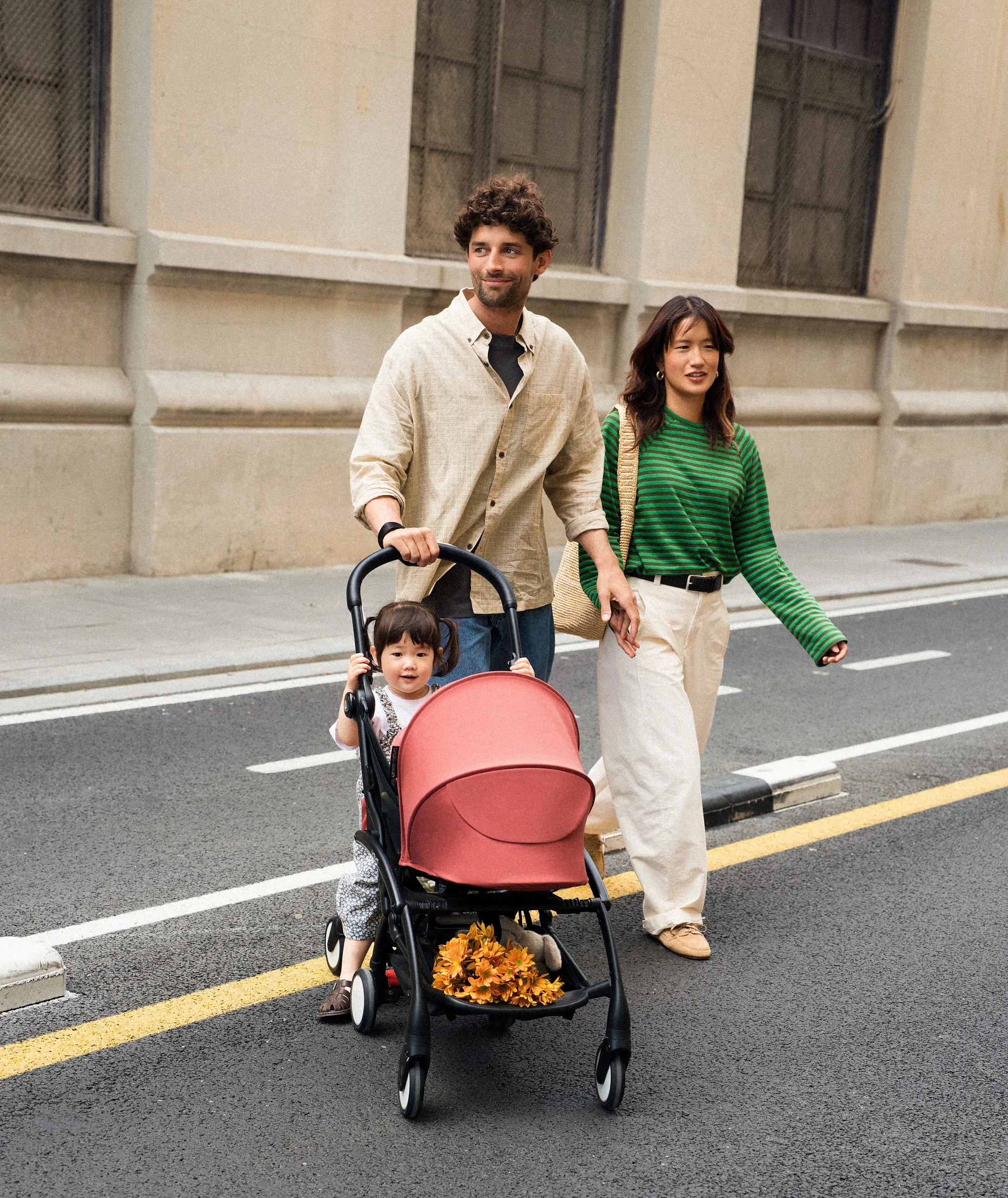 A lifestyle shot of a woman pushing a Stokke stroller with a child inside on a sunny tree-lined urban street.