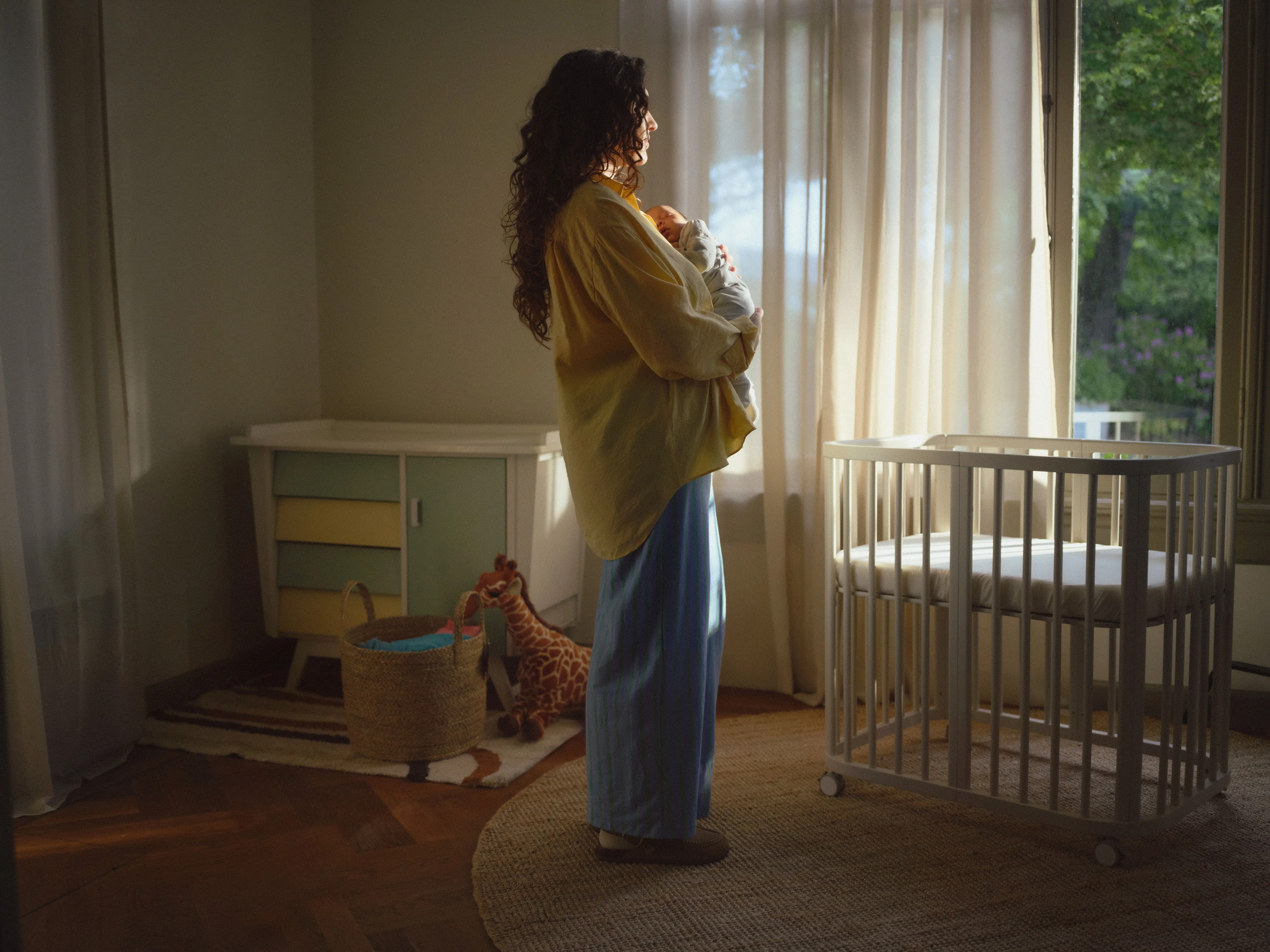A peaceful baby sleeps in a Stokke Sleepi™ oval wooden crib in a softly lit nursery setting.