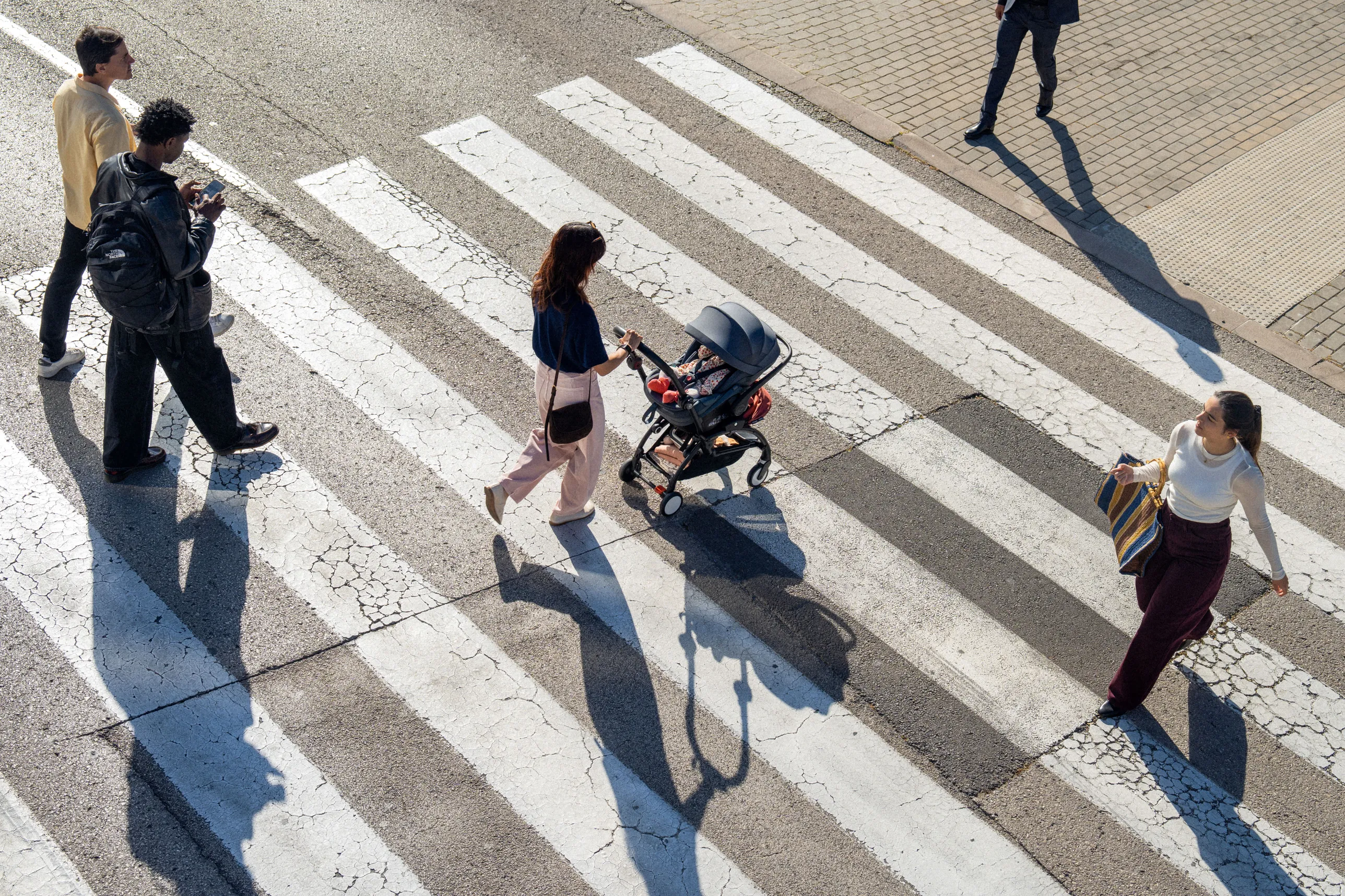 A lifestyle shot of a woman carrying an infant in a black baby carrier while pushing a stroller on a brick-lined urban street.