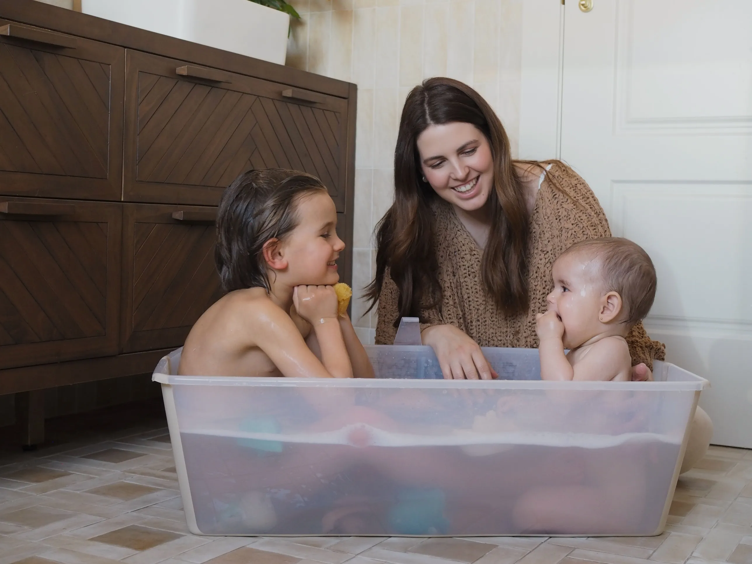 A lifestyle shot of a toddler in a bathtub being bathed by a caregiver, holding a green bathing cup in a bright, clean bathroom setting.