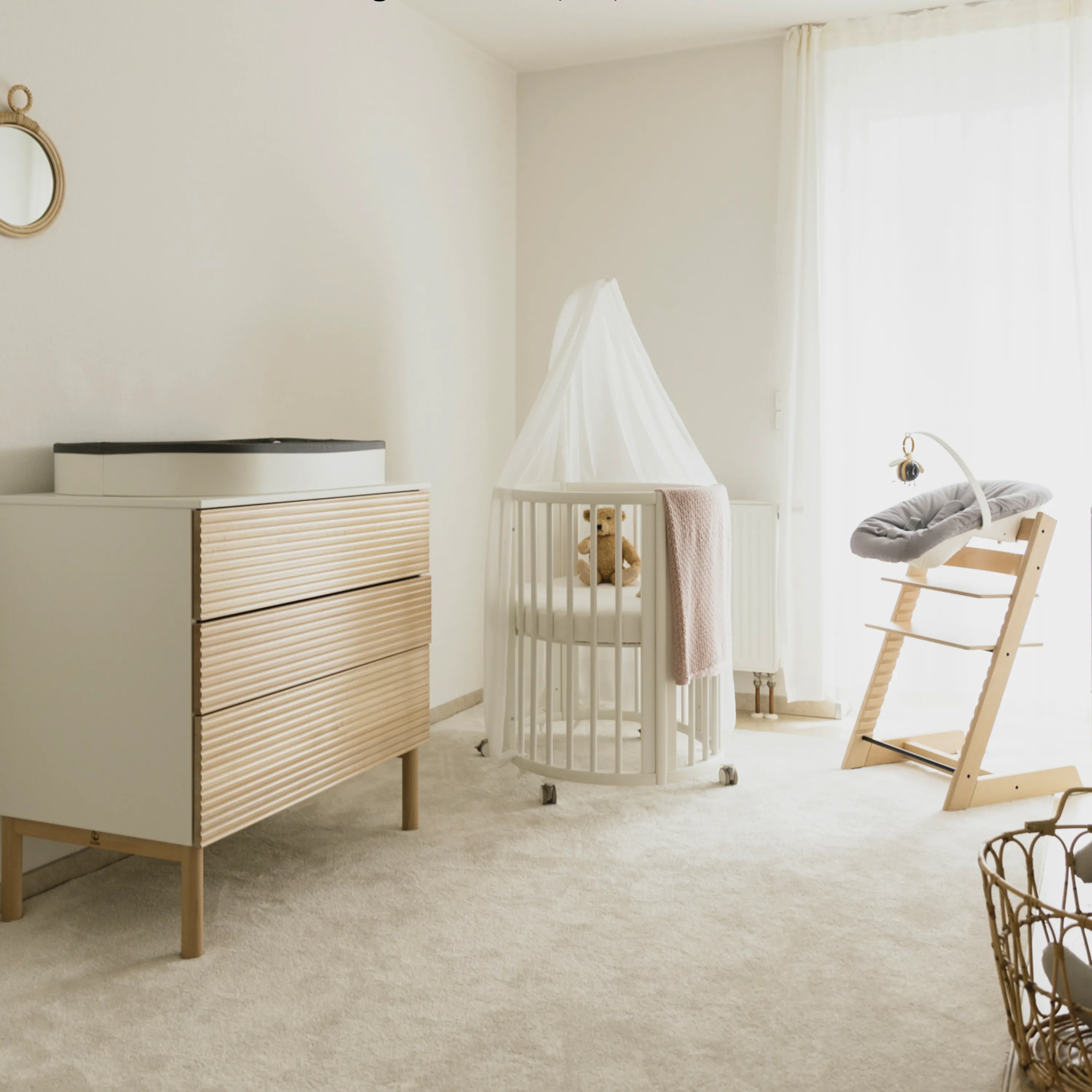 A lifestyle shot of the Stokke® Nighti™ Mini Newborn Cot in a well-organised small nursery, showing its compact space-efficient shape, slim frame and wheels in a calm, airy setting.