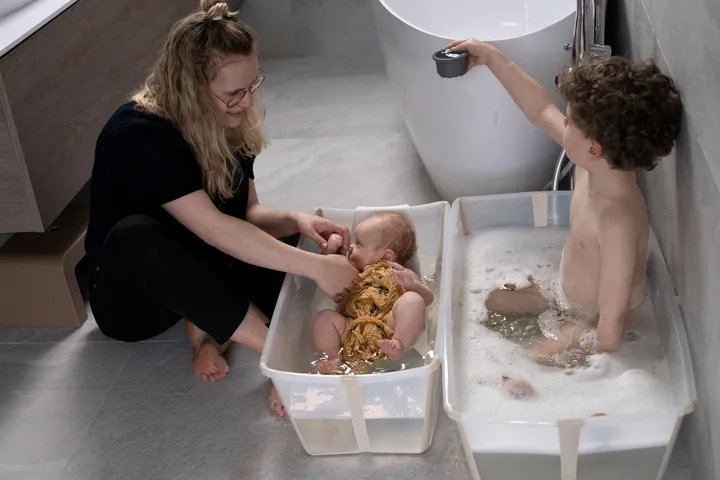 A lifestyle shot of a man and toddler bathing together in a light blue portable bath basin in a modern concrete nursery setting.